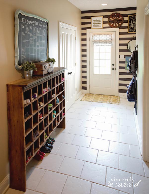 Rustic shoe presentation. This is a functional mud room, with stripe wallpaper, vintage shoe cabinet, chalkboard, white doors. The Designer creatively added the strips and made the chalkboard calendar from an old mirror. And this infuses rustic look to this stylish space. 